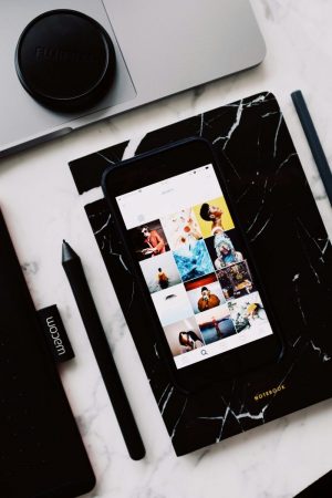 A smartphone displaying a grid-style photo gallery lies on a black marble notebook. Nearby, there’s a closed laptop with a camera lens cap, a stylus, and a Wacom tablet pen resting on a white marble surface.