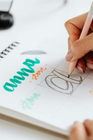 A person practices hand lettering in a sketchbook, writing the word “anna” in colorful inks and sketching a letter “a.” Their hand holds a pen, and color swatches and a clipboard are visible on the white desk.